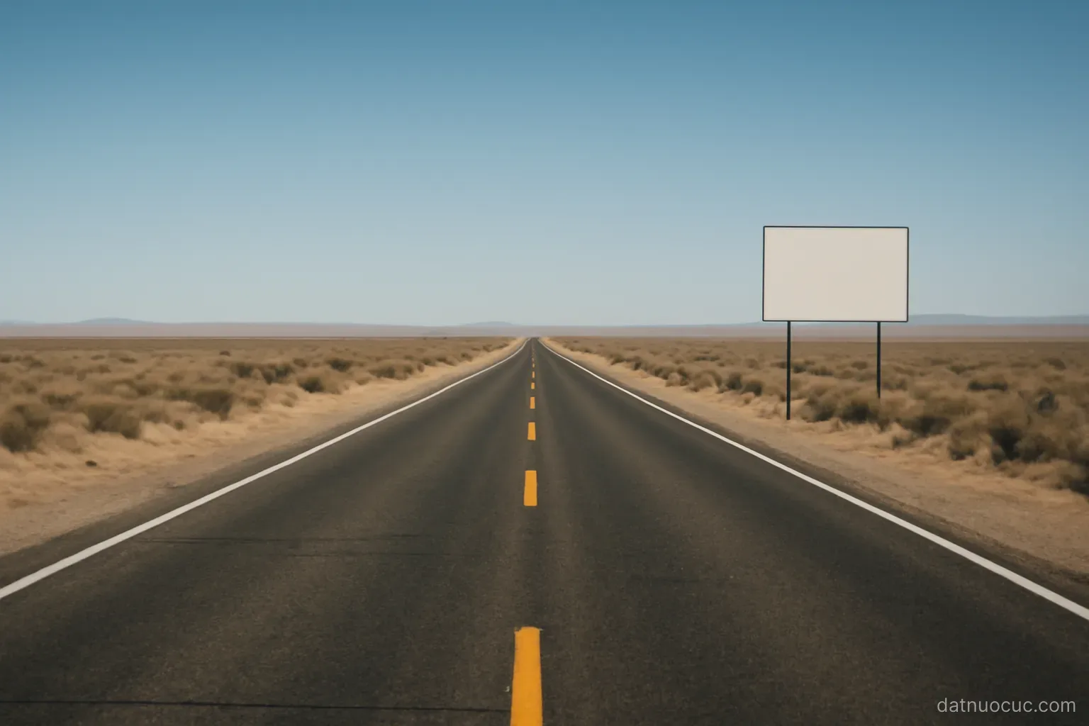 An empty highway cutting through flat dry scrubland toward the horizon, clear sky, no vehicles