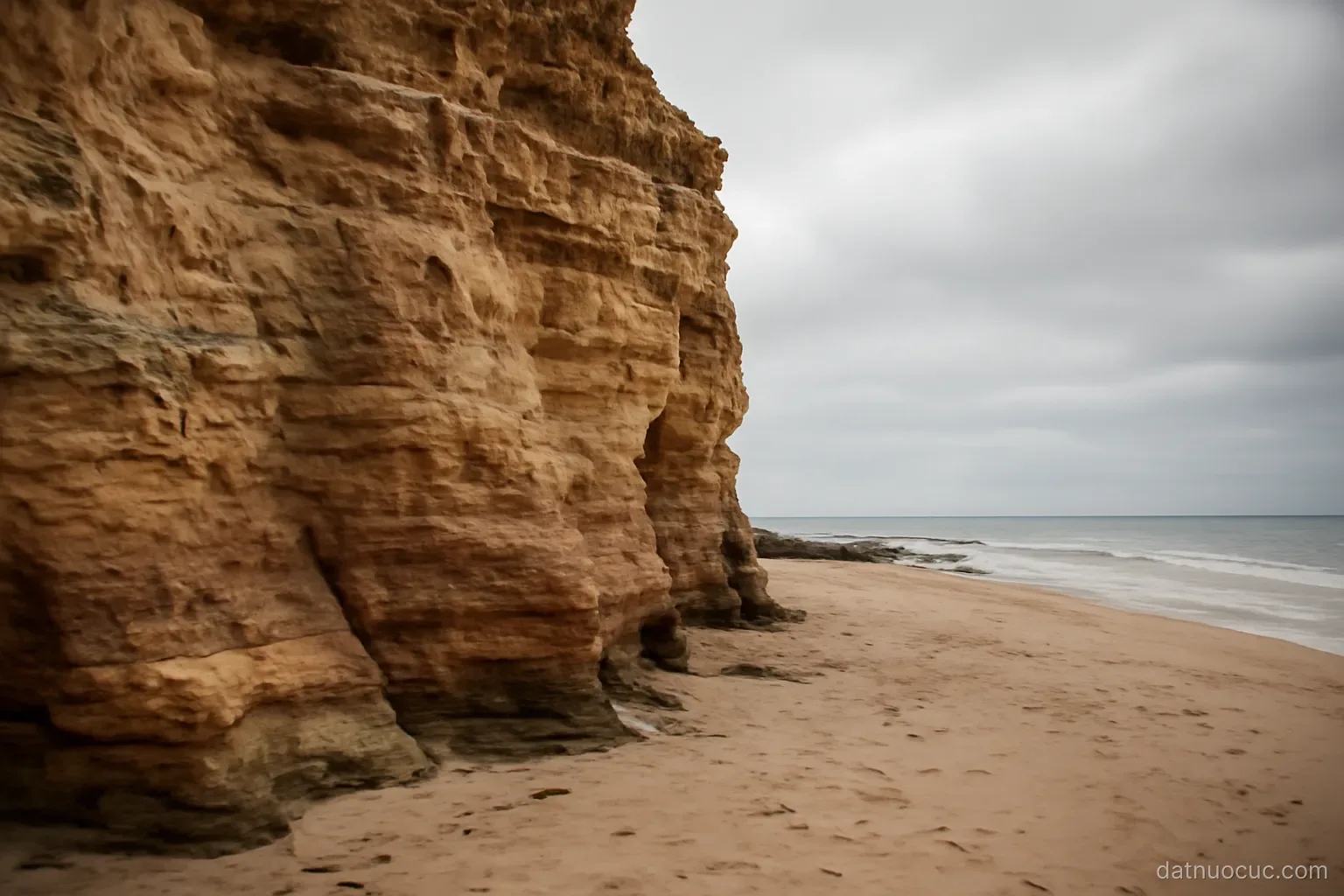 A weathered sandstone cliff face beside an empty beach at low tide, overcast sky, no people
