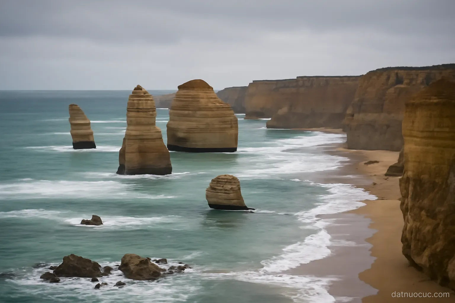 Tall limestone sea stacks rising from the ocean beside a cliff coastline, overcast sky, no people